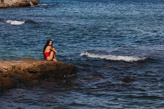 A woman in a vibrant red swimsuit sits gracefully on a rocky edge, surrounded by the endless blue of the Mediterranean Sea, embodying the spirit of freedom, elegance, and slow beauty.